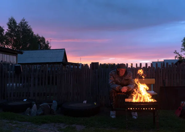 傍晚时分 一位老人一个人坐在村子里的长椅上 坐在篱笆后面 凝视着炉边的炉火 — 图库照片