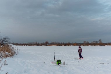 Kışın balıkçı kız, elinde oltayla buzun üzerindeki kutuda yürüyor. Gölün kenarındaki buz kırıcının yanında akşam gökyüzünün arka planında ve ufukta ağaçlarla..