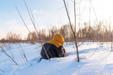 Sırtında kapüşonlu bir adam kışın çalıların arasında bir kar yığınına tırmanıyor..