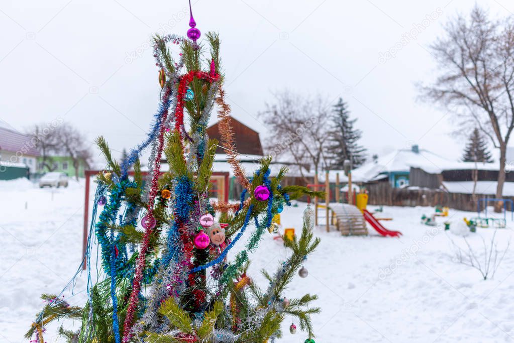 A Christmas tree stands decorated with garlands and tinsel in a snowdrift on a street in the Russian city in a courtyard with children's slides with private cottages for the new year holiday.