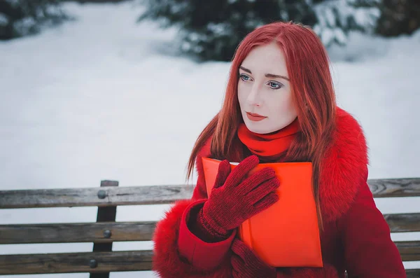 Mujer joven con pelo rojo en abrigo rojo en el bosque de invierno con un libro — Foto de Stock