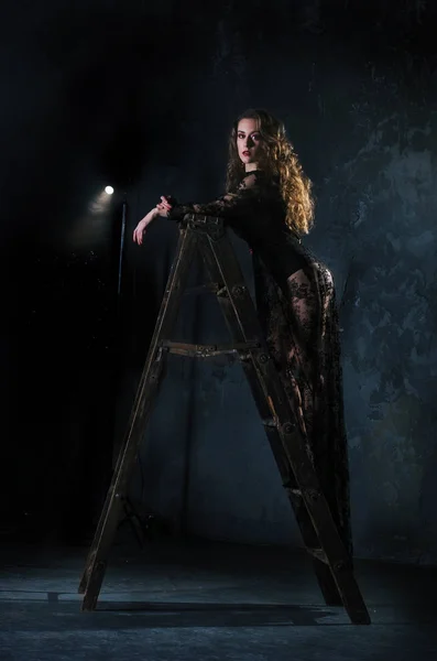 Eine attraktive junge Frau in einem langen schwarzen Kleid steht auf einer Leiter, einer Leiter gegen eine Grunge-Wand — Stockfoto