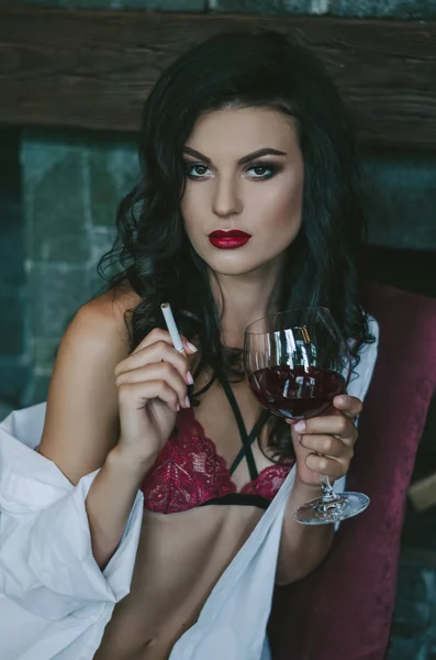 Portrait d'une brune en lingerie rouge et une chemise blanche. Elle fume une cigarette et tient un verre de vin rouge. Photo de mode — Photo