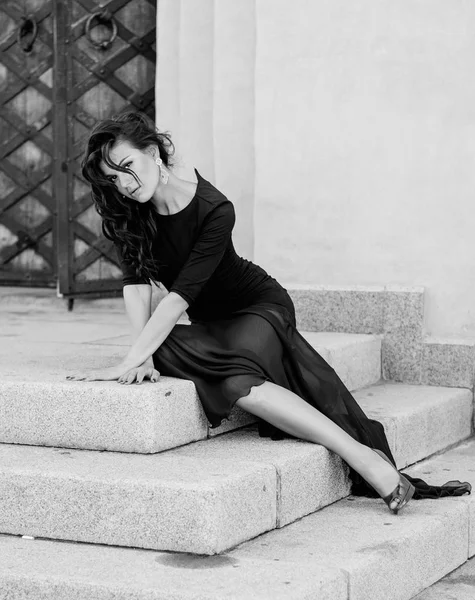 Eine attraktive sexy schlanke Brünette in einem langen schwarzen Kleid mit Schlitz sitzt auf den Stufen eines Vintage-Gebäudes. Modefoto. Schwarz-Weiß-Foto. — Stockfoto
