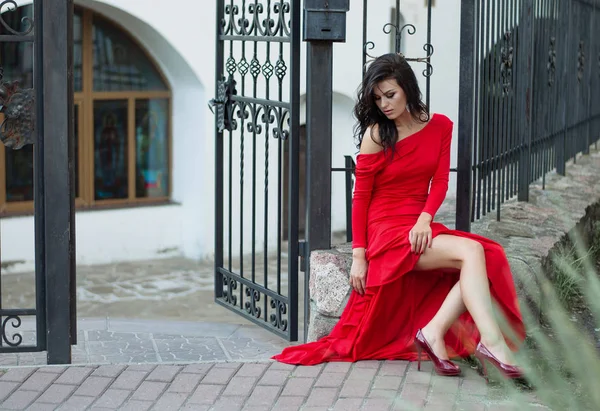 Una morena delgada y sensual con un largo vestido rojo se sienta a las puertas de hierro forjado. Foto de moda . — Foto de Stock