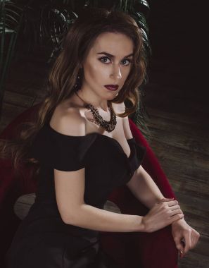 Kırmızı koltukta oturan uzun siyah bir akşam elbiseli bir çekici, seksi ince ince kahverengi saçlı kadın. Studio fotoğraf. Moda fotoğraf