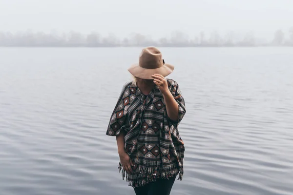 Mulher de chapéu e poncho em pé na margem do rio e olhando para a floresta de outono coberta com névoa matinal — Fotografia de Stock