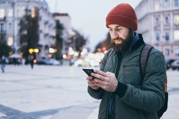 Hombre barbudo joven mirando en la pantalla del teléfono móvil mientras camina por la calle —  Fotos de Stock