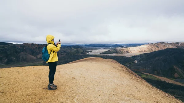 Woman in yellow raincoat hiking and taking pictures of the colourful mountains in Landmannalaugar national park, Iceland — 스톡 사진