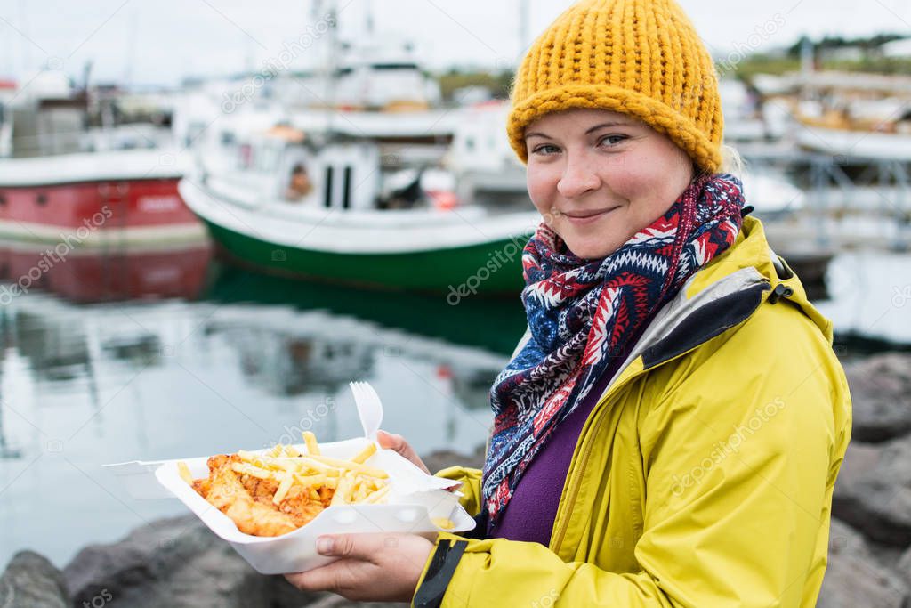 Young woman in yellow raincoat holds a portion of fish and chips in Husavik, Iceland