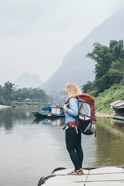 Kaukasische blonde Frau mit Rucksack steht auf Bootsanlegestelle im Dorf Muang Ngoi, Laos — Stockfoto