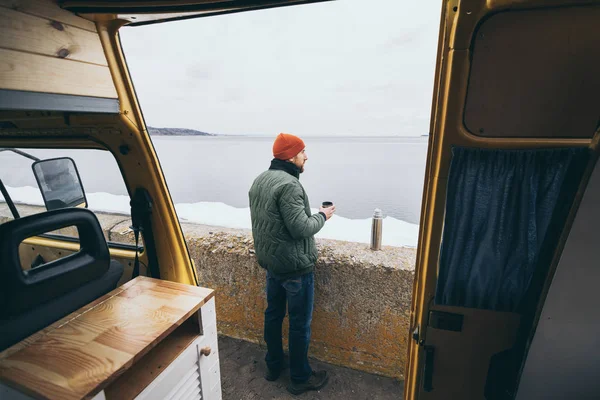 Junger Mann trinkt Tee aus Thermoskanne vor Wohnmobil mit See im Hintergrund — Stockfoto