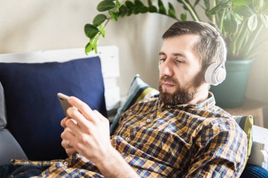Evdeki koltukta kulaklıkla müzik dinleyen bir adam cep telefonu ekranına bakıyor.