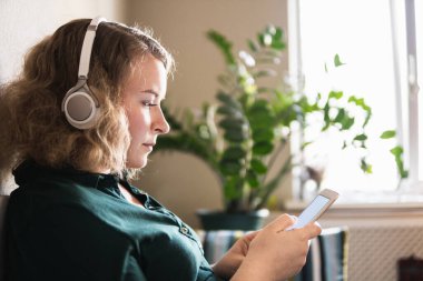 Genç sarışın kadın evdeki koltukta kulaklıkla müzik dinliyor, cep telefonu ekranına bakıyor.