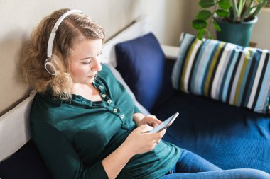 Genç sarışın kadın evdeki koltukta kulaklıkla müzik dinliyor, cep telefonu ekranına bakıyor.