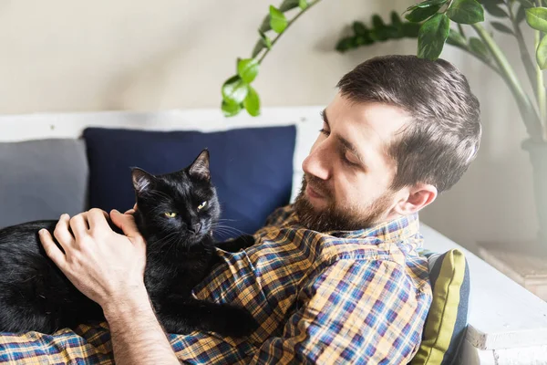 Man petting black cat while lying on the sofa at home, having a good time.