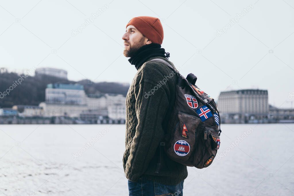 Young bearded traveller with patches on backpack standing at the river front with cityscape on background.