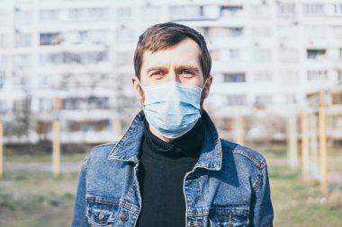 Arka planda ikametgah binası olan, dışarıda maske takan endişeli genç bir adam. Corona virüsü kavramsal görüntüyü karantinaya aldı.