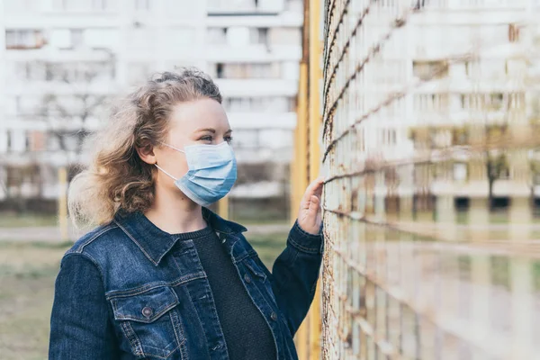 Dışarıda tıbbi maske takan Doğu Avrupalı bir kadın, arka planda ikametgah binası olan tel örgülerin yanında duruyor. Corona virüsü kavramsal görüntüyü karantinaya aldı.