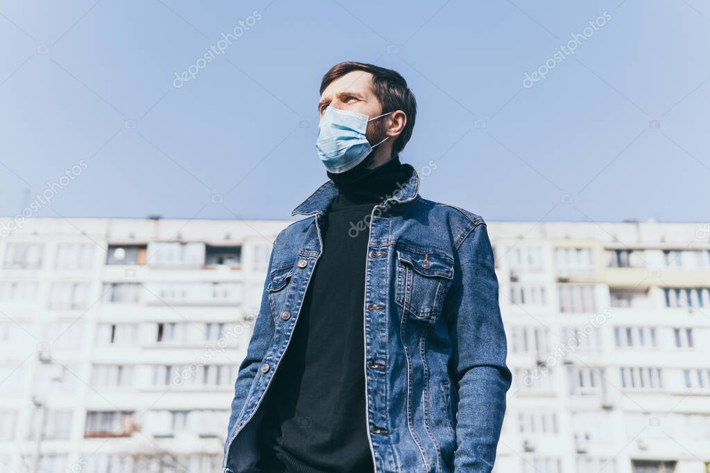 Concerned young man wearing medical mask outdoors with residential building on background. Corona virus quarantine conceptual image.