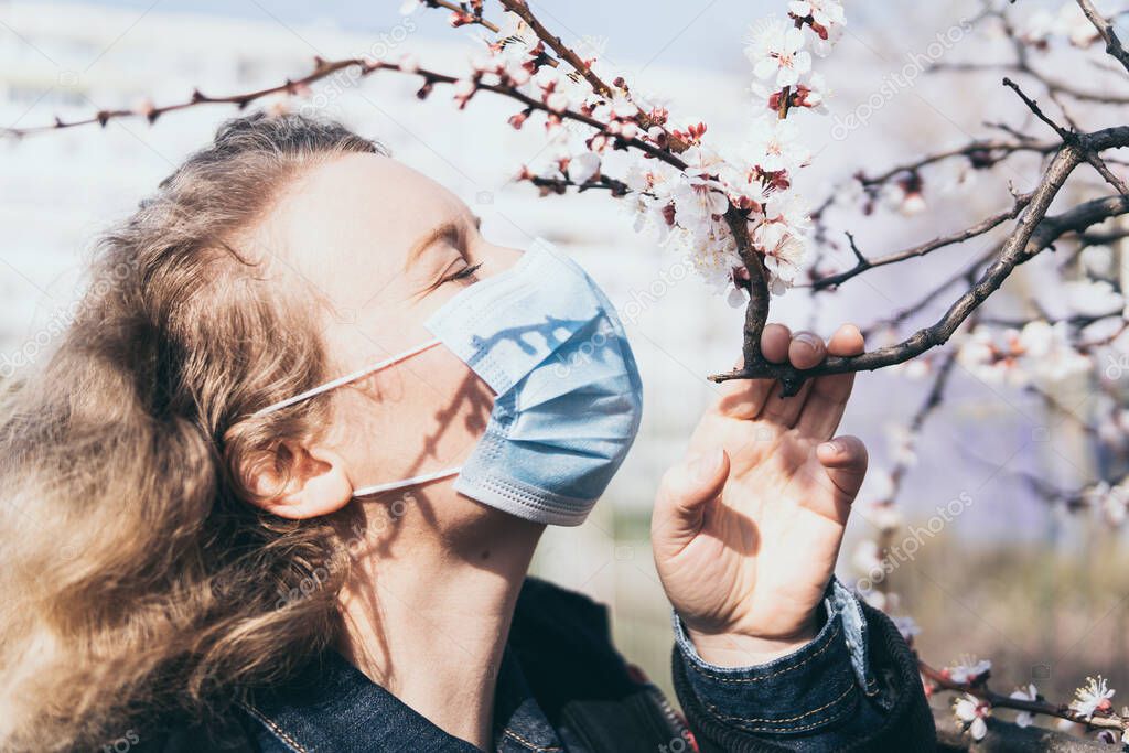 Young blonde woman smells cherry tree blossom through medical mask. Conceptual image of corona virus quarantine and allergy.