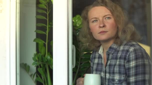 Junge blonde Frau, die mit besorgtem Gesichtsausdruck aus dem Fenster schaut. Während Coronavirus-Quarantäne zu Hause bleiben — Stockvideo