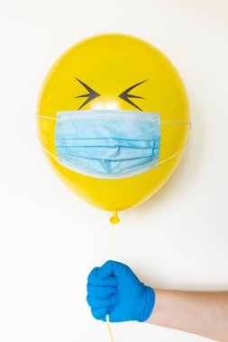 Hand in a rubber glove holding a yellow balloon with a medical mask and doodle closed eyes. Conceptual image of Birthday party during Corona virus quarantine lockdown clipart
