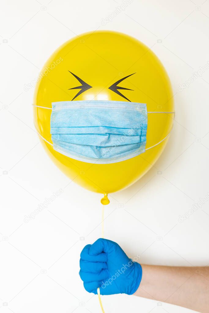 Hand in a rubber glove holding a yellow balloon with a medical mask and doodle closed eyes. Conceptual image of Birthday party during Corona virus quarantine lockdown