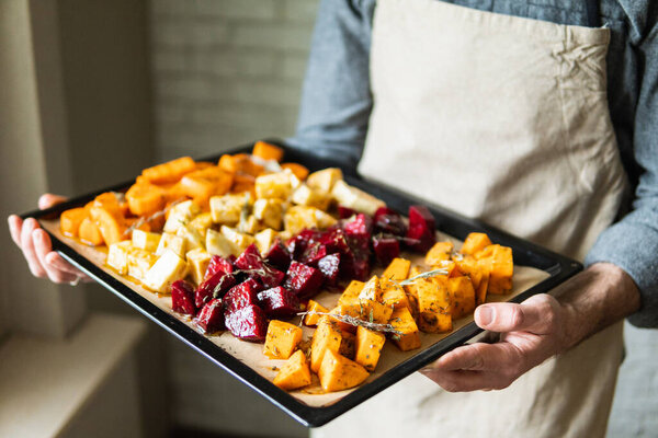 Male hands holding oven tray with vegetables mix. Vegetarian man cooking sweet potatoes and other veggies at home.