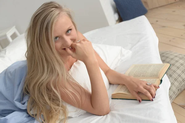 Красивая блондинка, лежащая на кровати комфортно мечтала и читала книгу. улыбаясь — стоковое фото