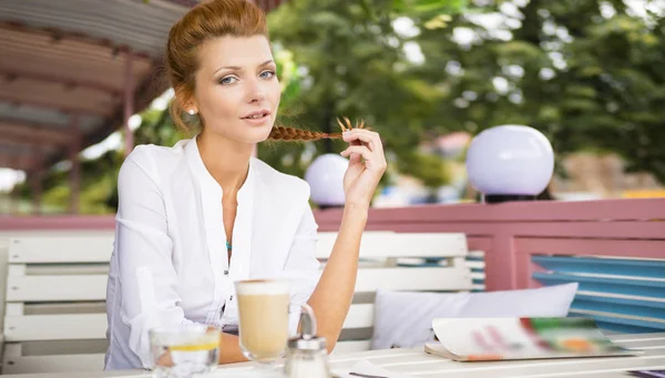 Mladý zasněný krásná žena sedí v letní kavárně, shlíží na ulici a pije horká káva latte z poháru. Royalty Free Stock Obrázky