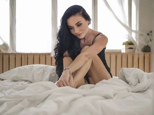 Close-up de sexy jovem mulher em lingerie posando na cama com belos olhos cinzentos. Menina morena com roupa interior preta em seu quarto — Fotografia de Stock