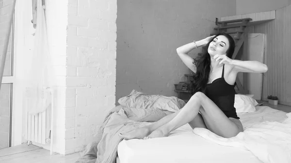 Привлекательная и расслабленная соблазнительная молодая женщина с распущенными волосами и стройным телом в нижнем белье лежит на белых простынях в спальне рано утром. Черно-белое изображение . — стоковое фото