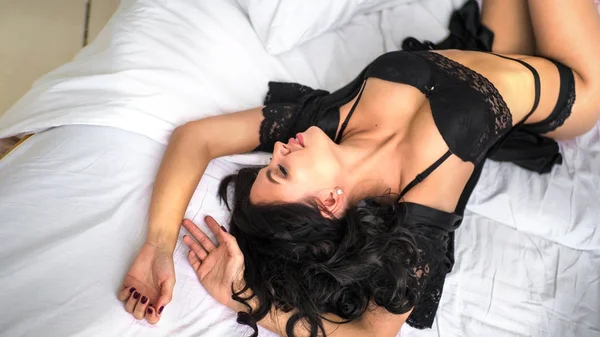 Sexy hermosa mujer morena tumbada en la cama en lencería negra sensual —  Fotos de Stock