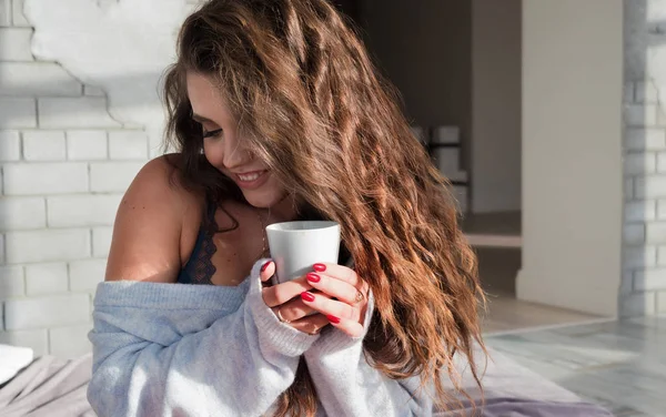 Chica sexy por la mañana con una taza de café en la cama en lencería y suéter. Cabello rizado, mujer rusa sosteniendo una taza con luz natural junto a la ventana en una habitación de matrimonio blanca . — Foto de Stock