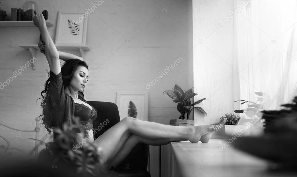 Sensual woman sits next the window in lingerie in her apartments early in the morning. Black ang white picture. Old hollywood style. She woke up and stretched in her chair