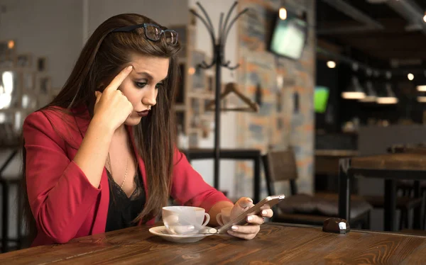 sad young woman looking at the screen of her phone during her coffee break. Beautiful woman working by the window in the cafe using her phone