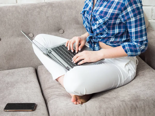 A mulher está a trabalhar sentada no sofá. As mãos estão segurando o telefone celular e trabalhando com um notebook . — Fotografia de Stock