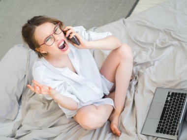 Dizüstü bilgisayar ile evde yatakta oturup sırasında cep telefonu tutan deli bir genç kadın portresi