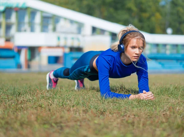 Młoda fitness lady w słuchawki zrobić deska zewnątrz. — Zdjęcie stockowe