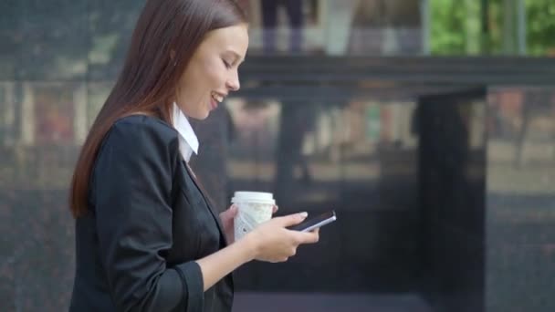 Donna in giacca e cravatta che cammina per strada con una tazza di caffè da asporto in mano e messaggiando al telefono — Video Stock