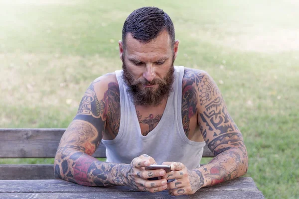 纹身有胡子的人用智能手机，在公园里写 — 图库照片