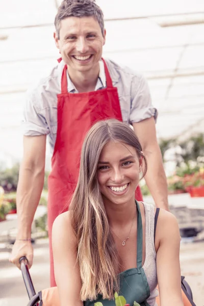 Sellers couple have fun pushing the wheelbarrow in a greenhouse — Stock Photo, Image