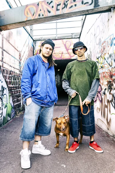 Dos cantantes de rap con un perro en un metro con graffiti en la parte de atrás — Foto de Stock