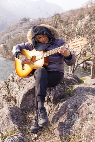 Belle femme mature jouant une guitare assise sur un rocher — Photo