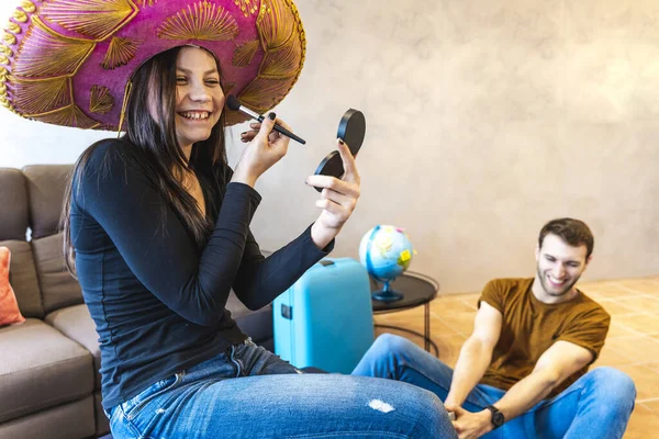 young womanwearing sombrero is finishing her make up before leaving for the vacations and her boyfriend is impatient to leave. funny holidays concept
