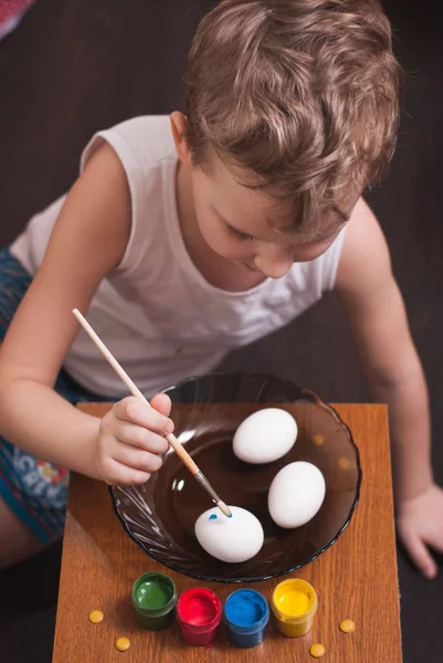 Un petit garçon peint les œufs à la veille de Pâques, peintures colorées et brosse , — Photo