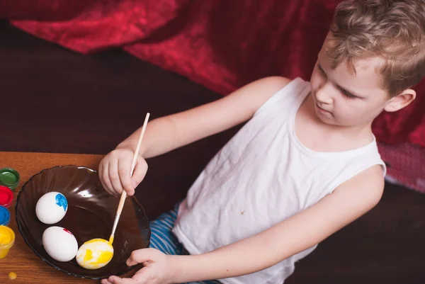 Un petit garçon peint les œufs à la veille de Pâques, peintures colorées et brosse , — Photo