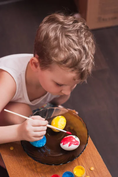 Un petit garçon peint les œufs à la veille de Pâques, peintures colorées et brosse , — Photo