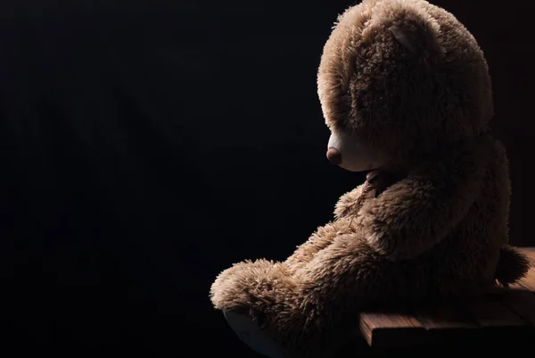 A lone Teddy bear sitting in the dark, side view, — Stock Photo, Image